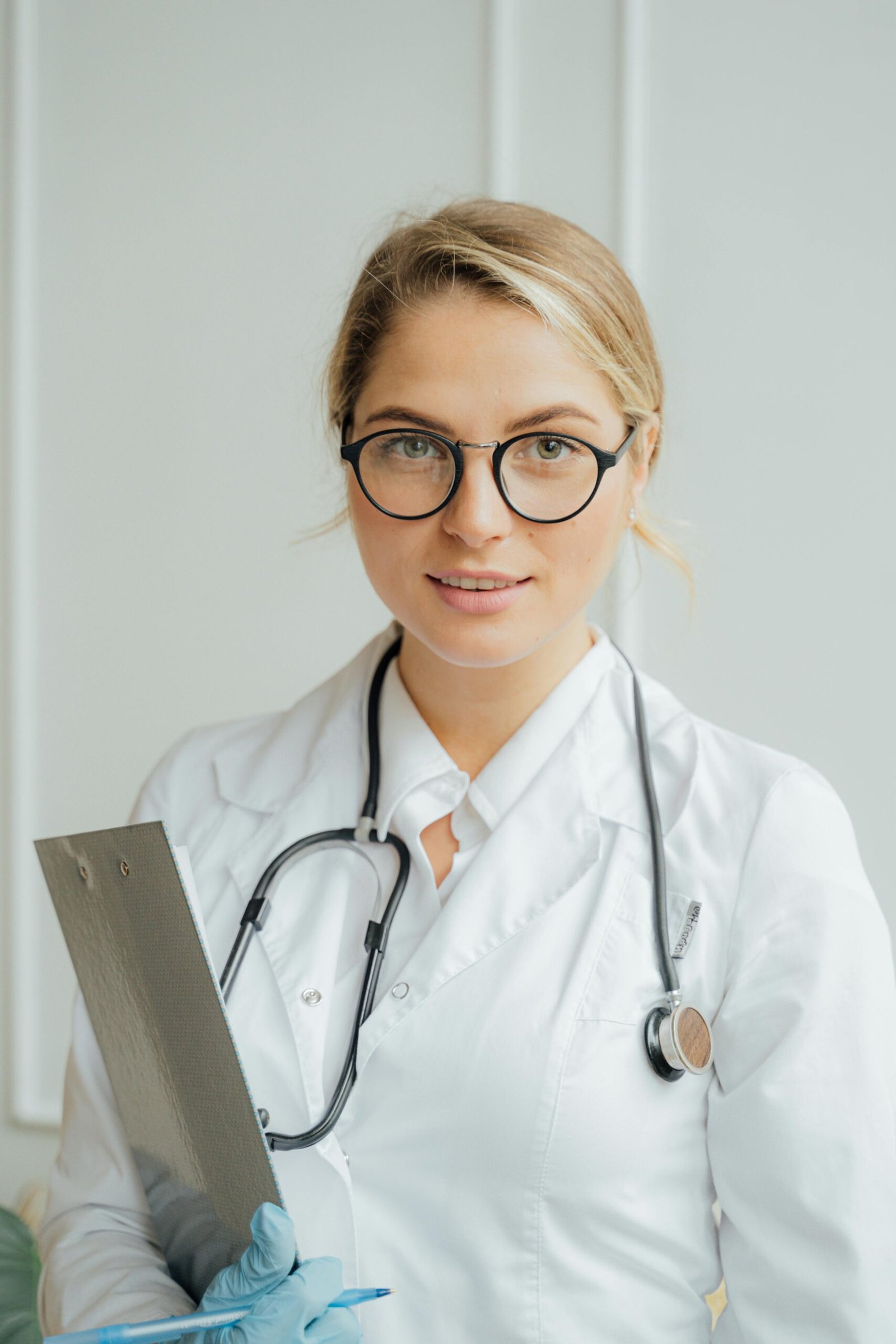 About Us Smiling female healthcare professional in white coat holding clipboard indoors.